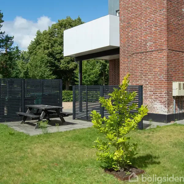 En sort picnicbænk på en flisebelagt terrasse støder op til et murstenshus. En grøn busk og plæne omgiver området med sorte hegn i baggrunden. Træer ses længere væk.