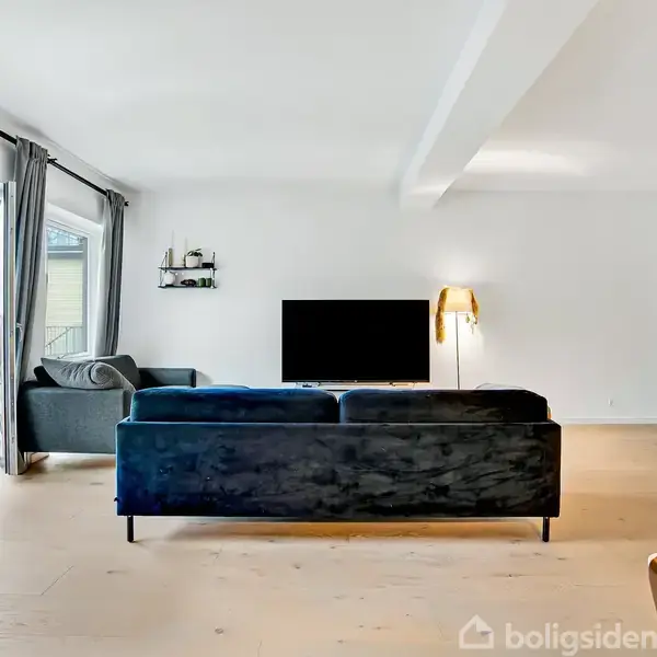 Sort sofa vendt mod en vægmonteret fladskærm i en lys stue. Store vinduer med mørke gardiner giver udsigt til en altan. En væglampe lyser rummet.