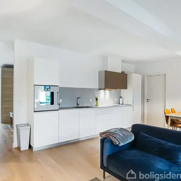 En moderne stue viser en blå sofa foran et åbent køkken med hvide skabe. Til højre er et spisebord med fire stole og en lampe i loftet.