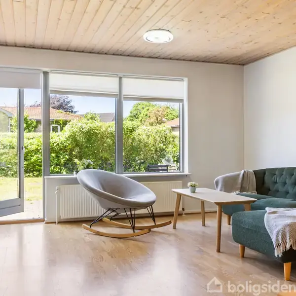 Stue med en grøn sofa og gråt lænestol. Et sofabord står mellem dem. Lyst trægulv og træloft. Store vinduer åbne mod en frodig have. Ingen tekst i billedet.