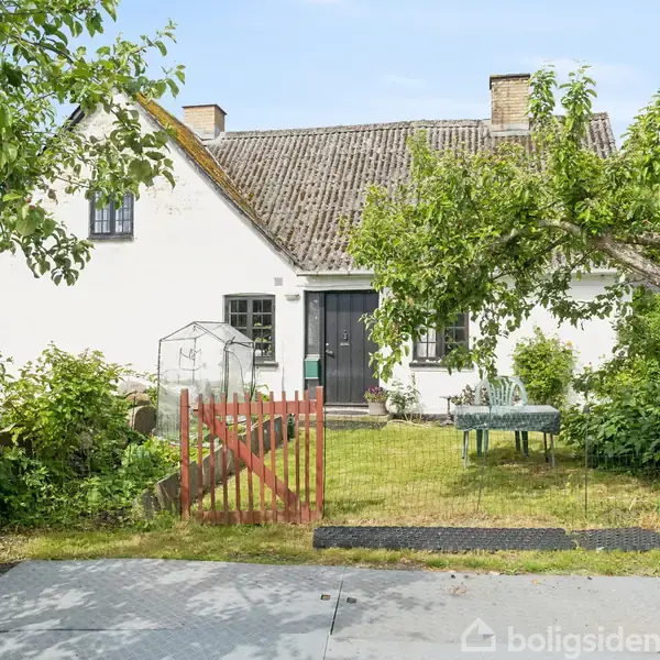 Et hvidt hus med et skifertag står omgivet af en frodig have med træer. En rød låge fører til en grusbelagt sti, der fører til hoveddøren.