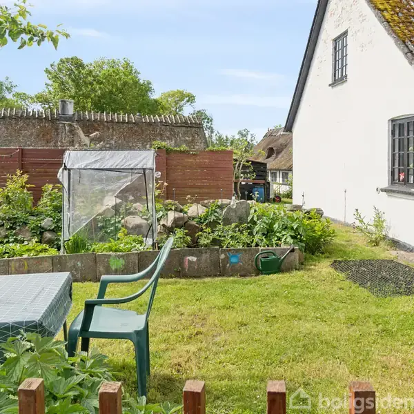 Hvide hus står ved siden af en have med bord og stol. Drivhus og planter omringet af grøn beplantning og træhegn. Blå himmel og træer i baggrunden.