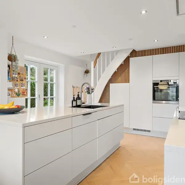 Et moderne køkken med hvide skabe og en central ø, hvorpå der står en frugtskål. En trappe ses i baggrunden, og dagslys strømmer ind gennem glasdøre.