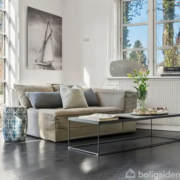 En sofa med puder står i en lys stue ved siden af et stort vindue. Et billede af en sejlbåd hænger på væggen. Et bord med blomster er foran sofaen.