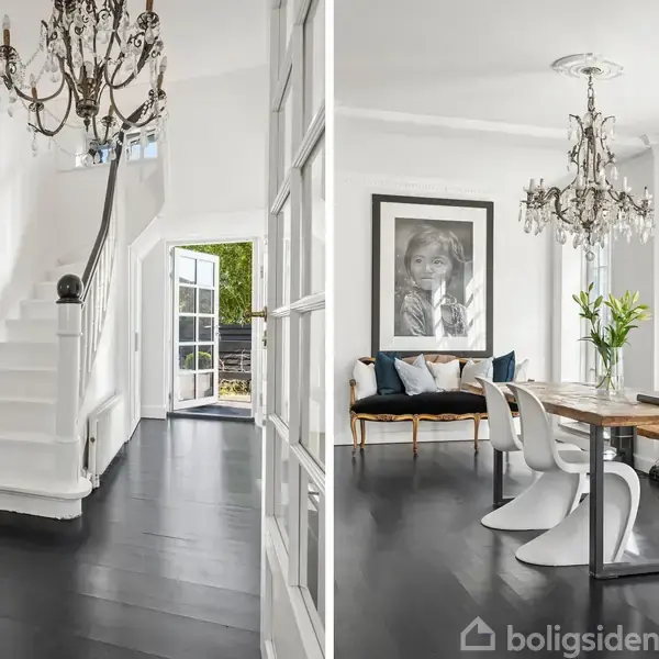 En lys og elegant gang med en hvid trappe til venstre. Spisestue med træbord og moderne hvide stole. Stor lysekrone hænger i begge rum, hvor sorte gulve og hvide vægge dominerer.