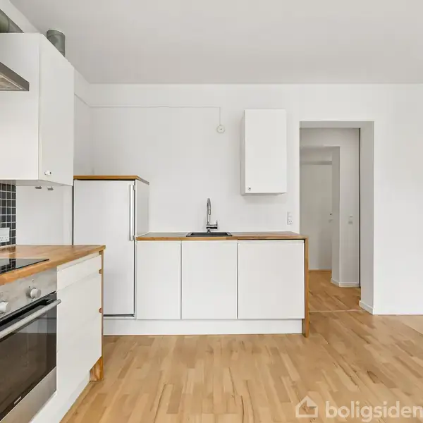 Køkken med hvide skabe og træbordplader, en indbygget ovn, og sort flisebelagt bagvæg, står ubenyttet. Rummet har lyst trægulv og hvide vægge, hvilket skaber en ren og moderne atmosfære.