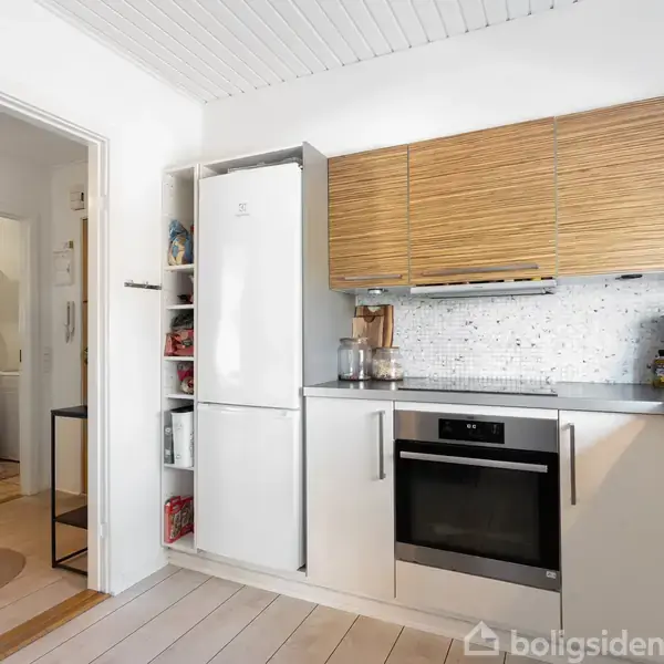 Køkken med hvidevarer, herunder køleskab, ovn og kaffemaskine, i moderne indretning. Lys træbeklædning og hvid mosaikvæg. Åben dør fører til tilstødende stue med runde tæpper.