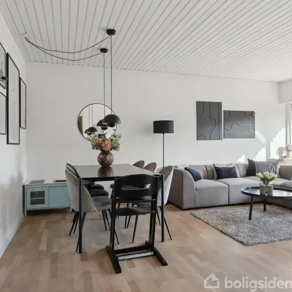 Spisebord med stole, placeret i lyse stue; ved siden af en hjørnesofa med sofabord på tæppe. Indretningen omfatter lamper, vægkunst og en vase med blomster.