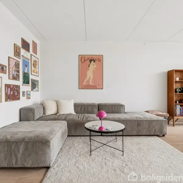 En stor, grå sofa står i en stue mod en hvid væg med mange billeder. Et sofabord med en lyserød vase står på et tæppe. En reol står i hjørnet.
