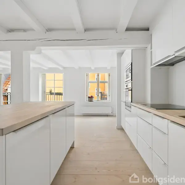 Køkken med hvide skabe og en stor ø disk med træoverflade, der vender ud mod vinduer i en lys stue. Vægge og loft er også hvide.