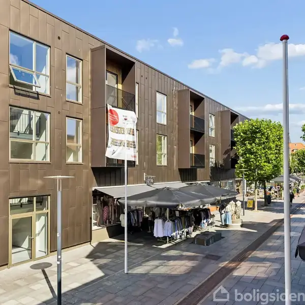 Bygning med moderne facade og store vinduer, dekorativt banner om boliger til salg hænger ned. Gade med fortovsbutik og træer i baggrunden, sollys og klar himmel skinner ned.