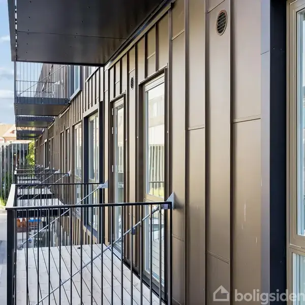 Altanrække med metalgelændere langs en moderne bygning, placeret over parkerede biler i urban omgivelser. Sollys reflekteres fra bygningen, og træer ses i baggrunden.