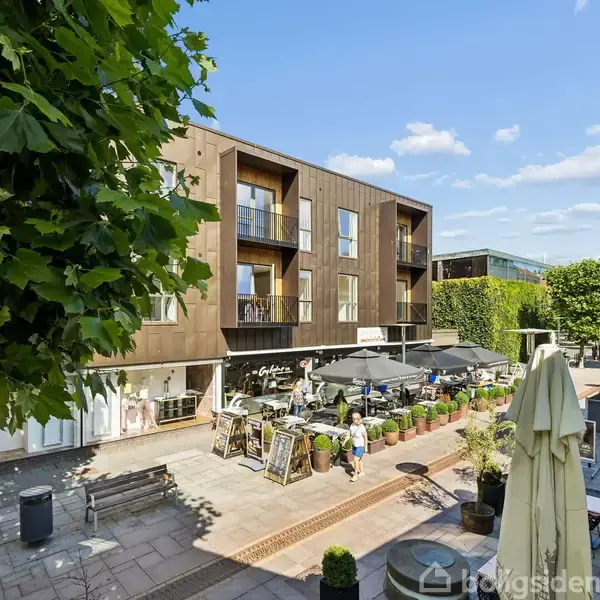 Et caféområde med udendørs siddepladser omgivet af planter foran en moderne bygning med træpaneler. Parasoller er slået op ved bordene, træer omkranser området under en blå himmel.
