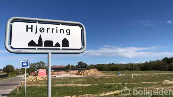 Skiltet med teksten "Hjørring" står ved vejkanten. I baggrunden er der en byggeplads og åbent landskab med blå himmel. En hastighedsbegrænsning på 40 km/t vises.