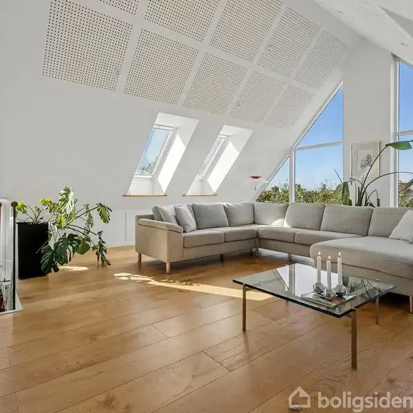 Stue med en stor grå hjørnesofa, stående på trægulv. Et glasbord med stearinlys foran sofaen. Grønne planter ved siden af. Sollys strømmer ind gennem skråvinduer.