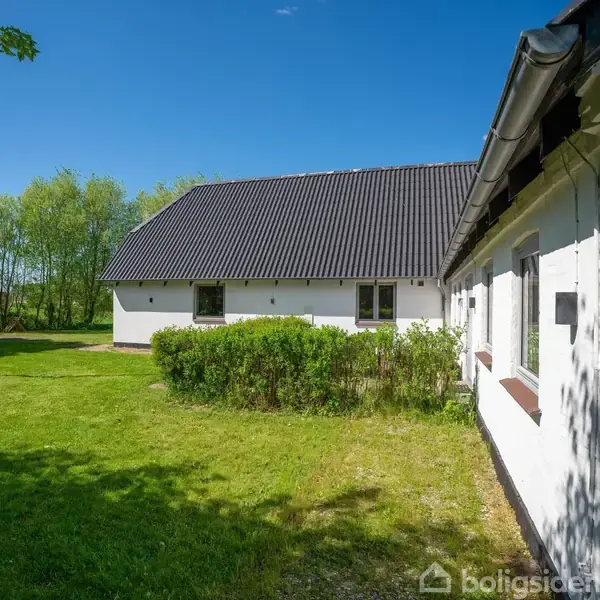 Hvidt hus med sort tag står omgivet af grønne træer og græsplæne i sollys; en flagstang ses ved siden af husets facade.