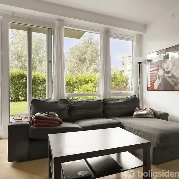 En grå hjørnesofa står foran store vinduer med udsigt til en grøn have. En plakat hænger på væggen, mens et sort sofabord står foran sofaen.