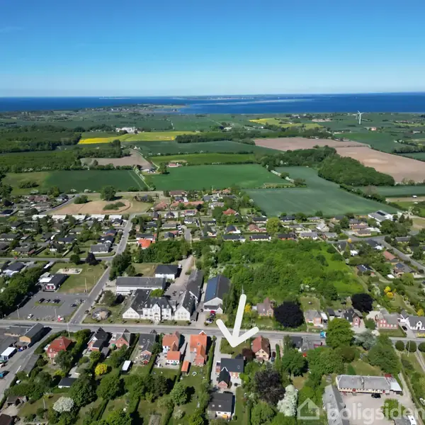 En pil peger på et hus i et beboelseskvarter med haver og veje. Omgivet af marker og havudsigt i baggrunden, under en klar blå himmel.