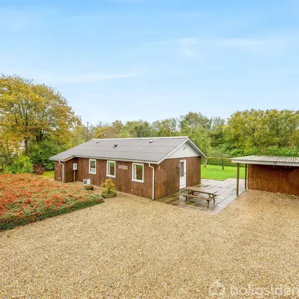 Et træhus står stille på en grusvej, omgivet af grønne træer og en lille rød busk. Ved siden af huset er en carport og et picnicbord.
