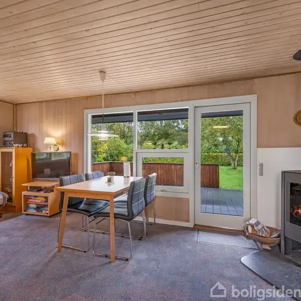 Et moderne stueområde med en tændt brændeovn i et hjørne. Foran står et spisebord med stole, omgivet af lyse trævægge og store vinduer med udsigt til en grøn have.