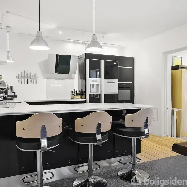 Køkkenindretning med sort køkkenø og tre barstole, kaffe- og espressomaskine på bordet. Hvide vægge, to hængelamper over bordet, og køkkenmaskiner inde i et skab, træbord i forgrunden.