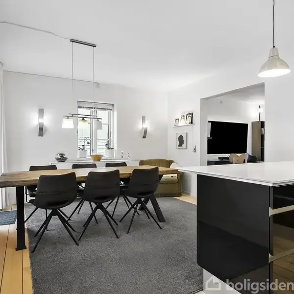 Spisebord med seks stole står på et tæppe i en stue, oplyst af loftlamper. Rummet har lyse vægge, store vinduer og en køkkenø med indbygget kaffemaskine i baggrunden.