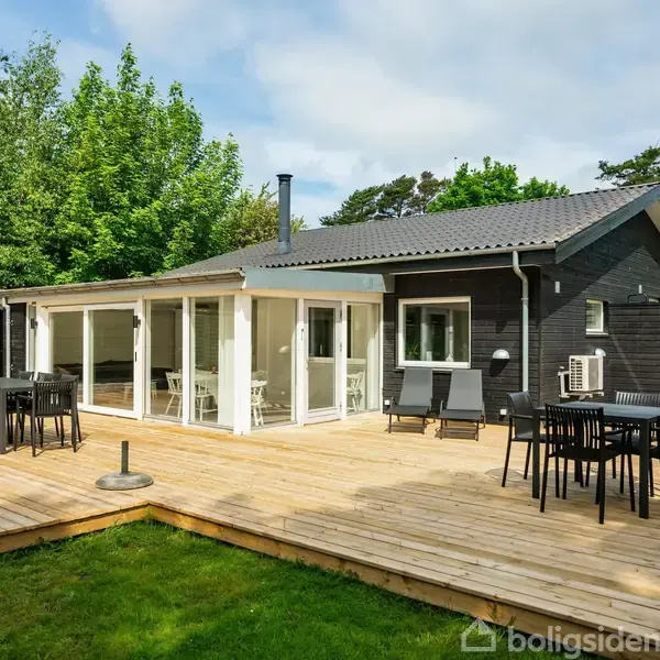 En moderne bungalow står på en stor træterrasse med borde og stole, omgivet af grønne træer og græsplæne, hvilket skaber en indbydende og afslappende atmosfære.