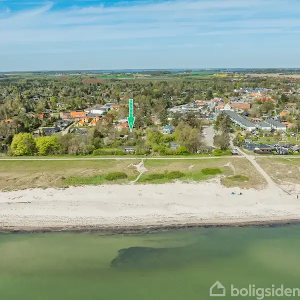 Strandkant strækker sig langs roligt vand, mens en by er synlig længere væk, omgivet af frodige træer. En grøn pil peger mod et område nær stranden.