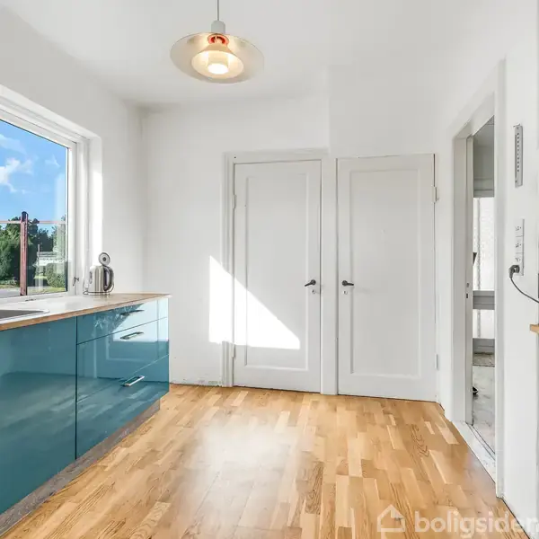 Køkken med blå skabe står ubenyttet, omgivet af trægulv og hvidmalede vægge. Sollys skinner gennem vinduet og belyser det sorte-hvide ternede fliseområde over bordpladen. Pejsø og mikroovn er synlige.