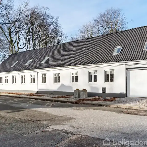 En lang, hvid bygning med skråt tag og flere vinduer står langs en vej. Omgivet af træer uden blade, skaber den en rolig atmosfære med lyse farvetoner.