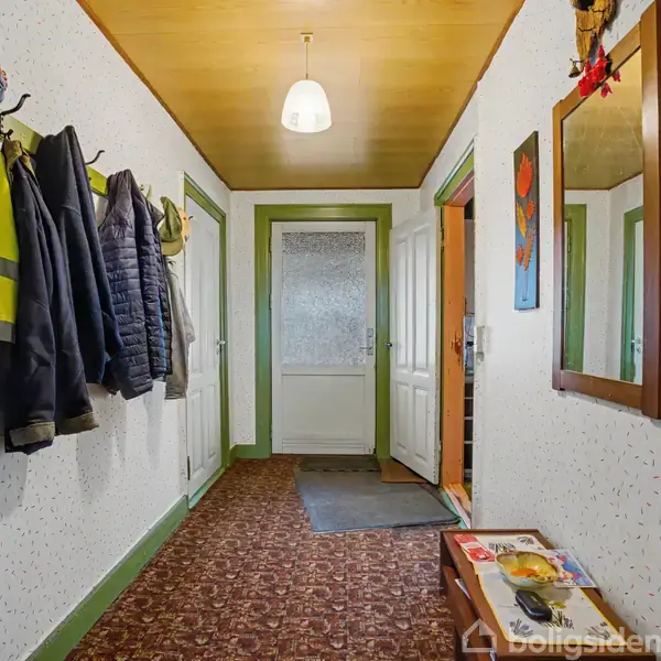 Gang med jakker hængende på knager, en lampe i loftet og et spejl på væggen. Dørene fører til andre rum. Der ligger en brochure på en skohylde.