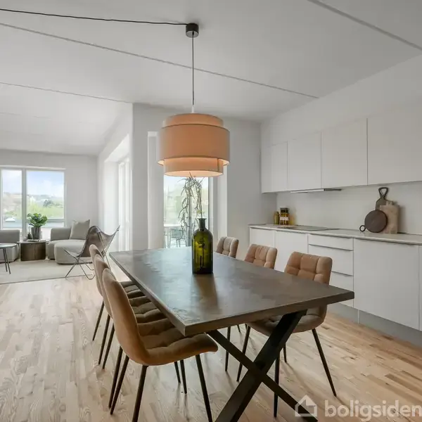 Et spisebord med seks beige stole står i et rummeligt, lyst køkken. En grøn flaske står på bordet. Køkkenet har hvide skabe og en bordplade i træ, med en udsigt til stue gennem store vinduer.