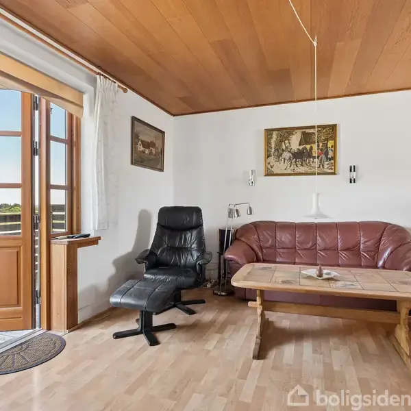 Stue med sofa og lænestol, placeret ved et rektangulært sofabord. En åben dør giver udsigt til natur. Væggen har malerier, og loftet er i træ.