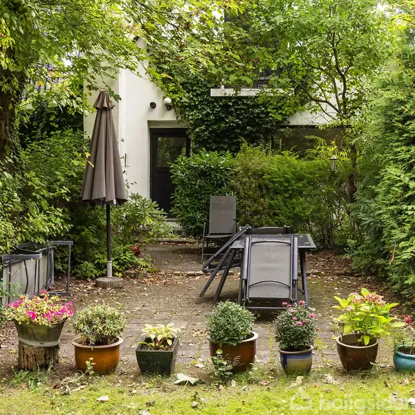 En terrasse omgivet af frodig vegetation, indeholder flere havestole og -borde. Rundt omkring står blomsterkrukker i forskellige farver. En parasol står til venstre for stolene.
