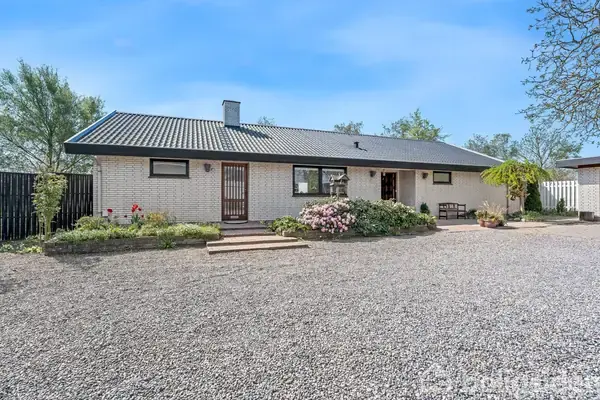 En en-etagers murstensbygning står i et grusbelagt område, omgivet af planter og buske foran huset. Træer ses i baggrunden under en blå himmel.