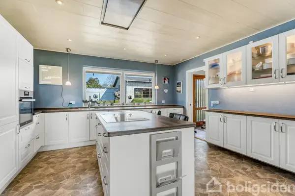 Køkken med hvidt skab og ovn til venstre, en stor ø i midten og skabe med glaslåger til højre. Lys strømmer ind gennem et vindue i baggrunden.