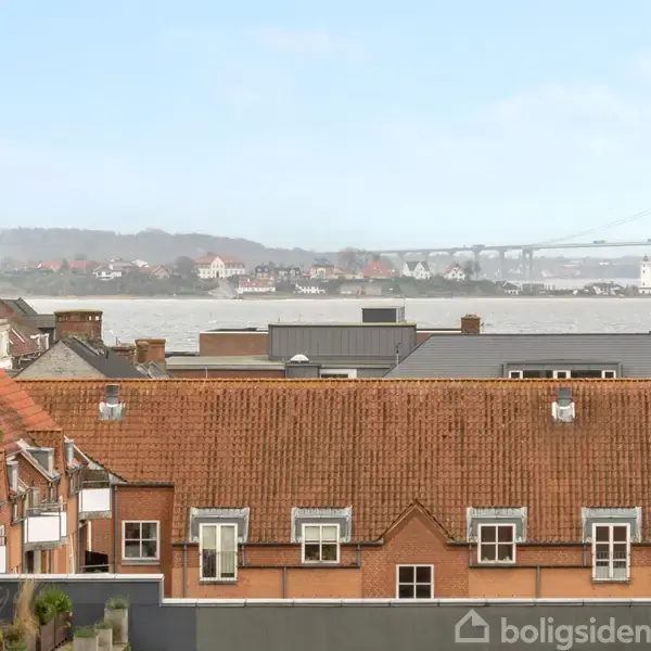 Bygninger med røde tegltage ses fra en højere vinkel, i baggrunden strækker en bro sig over vandet under en lyseblå himmel.