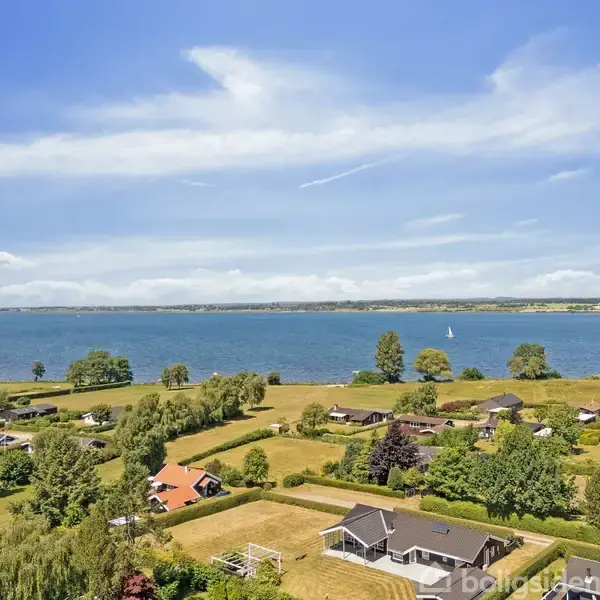Huse ligger spredt blandt grønne marker med en stor sø i baggrunden. En sejlbåd er synlig på vandet under en klar blå himmel.