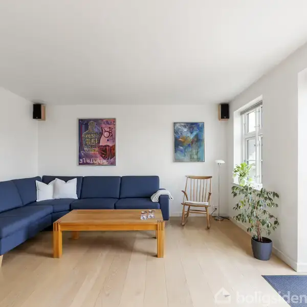 Blå sofa står stille i en lys stue med trægulv. Ved siden af er et træbord. Der hænger to malerier over sofaen, og en dør til højre åbner ud til en have.