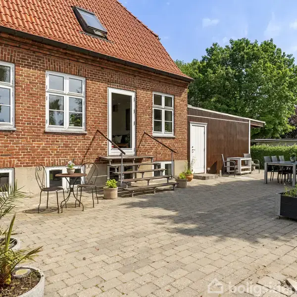 Rødt murstenshus med tag i tegl, trapper leder til terrasse med borde og stole, omgivet af planter og træer under en blå himmel.