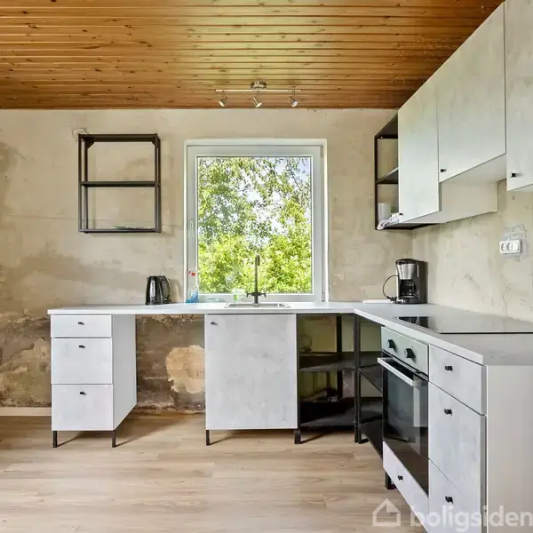 Køkken med hvidt køleskab, komfur og skabe i minimalistisk design, står mod en rå væg. Trævæg i loftet. Vinduet viser udsigt til træer udenfor.