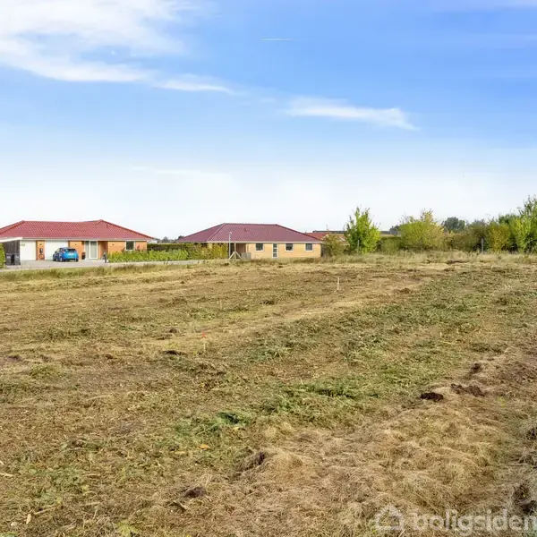 En tomt med kortklippet græs ligger i forgrunden. I baggrunden ses flere huse med røde tage. Området er omringet af træer under en blå himmel.
