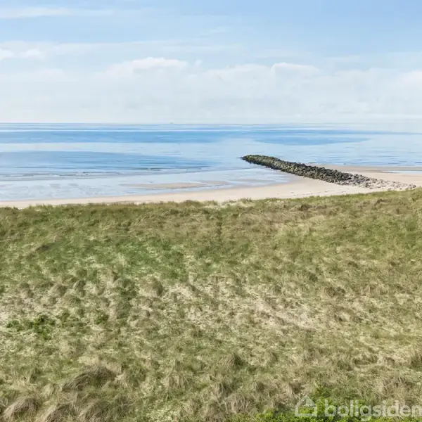 Græsareal strækker sig ud mod en stille kystlinje, hvor en stenvold beskytter mod havet. Vand og skyfri himmel mødes i horisonten, der skaber en rolig atmosfære.