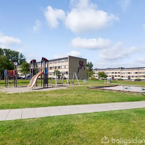 Legeplads med rutsjebane og gynger står på græsplæne foran moderne, lange lejlighedsbygninger. Sandkasse ses til højre, og grønne træer omgiver området under en skyet himmel.