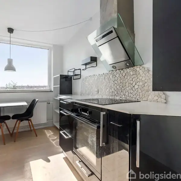 Et moderne køkken med sorte skabe og en ovn under en emhætte, står i et lyst rum med stort vindue og et bord med to stole.