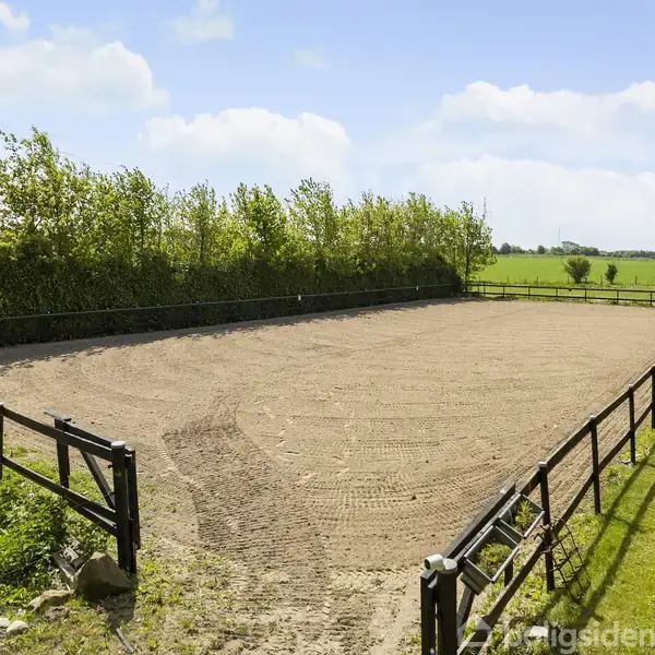 Sandet ridebane er omkranset af et lavt hegn; den står tom i landlige omgivelser med grønne marker og buske i baggrunden. Himmel med enkelte skyer ses øverst.