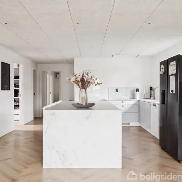Et lyst køkken med hvidt køkkenbord, moderne grå skabe og sorte hvidevarer. En buket tørrede blomster står på bordet. Trægulvet har sildebensmønster, og en hjortetrofæ hænger på væggen.