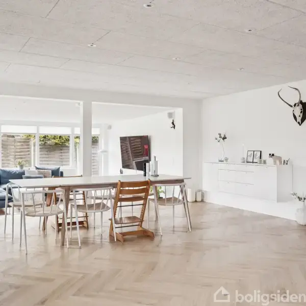 Spisestue med langbord og stole, indrammet af hvidmalede vægge og lyst parketgulv. I baggrunden ses en dagligstue med sofa og store vinduer, som lukker naturligt lys ind.