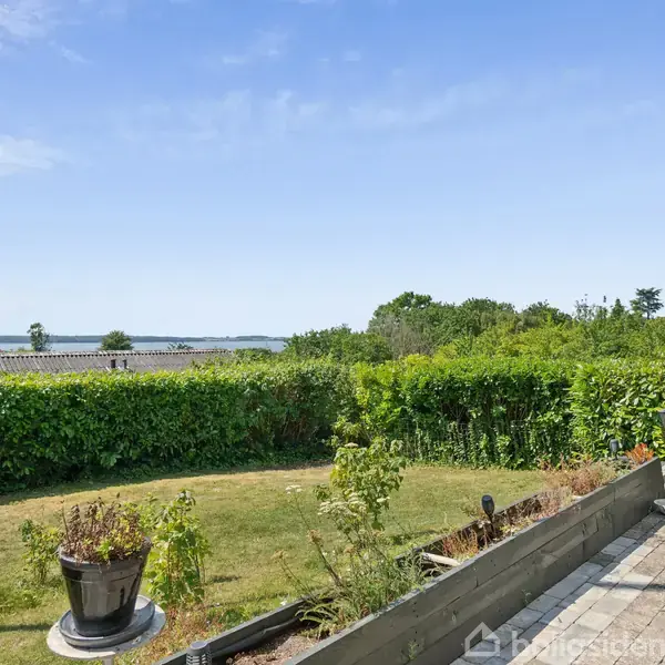 En have med græsplæne i forgrunden, prydet med blomsterkrukker. I baggrunden ses tætklippede hække og udsigt over et fjordlandskab, der omfavnes af klar blå himmel.