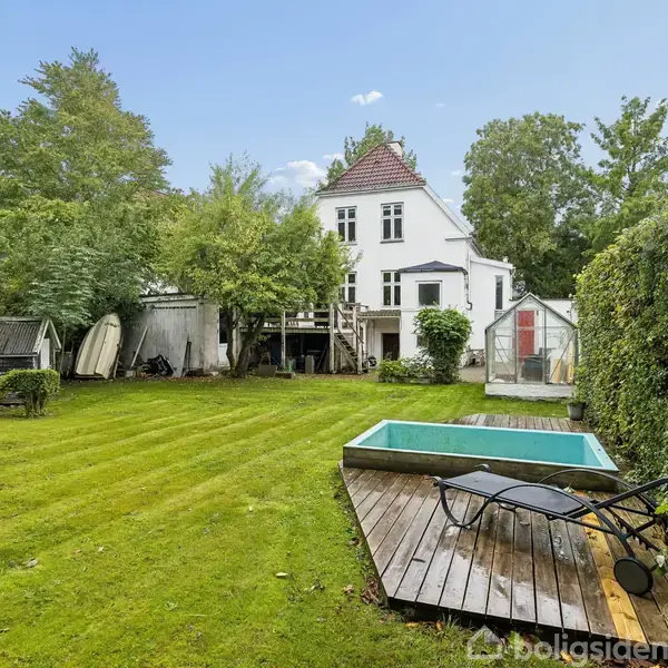Hus med hvid facade set fra baghaven. Der er en pool indrammet af træbeklædning og en liggestol på terrassen. Haven er omgivet af tæt beplantning og træer.